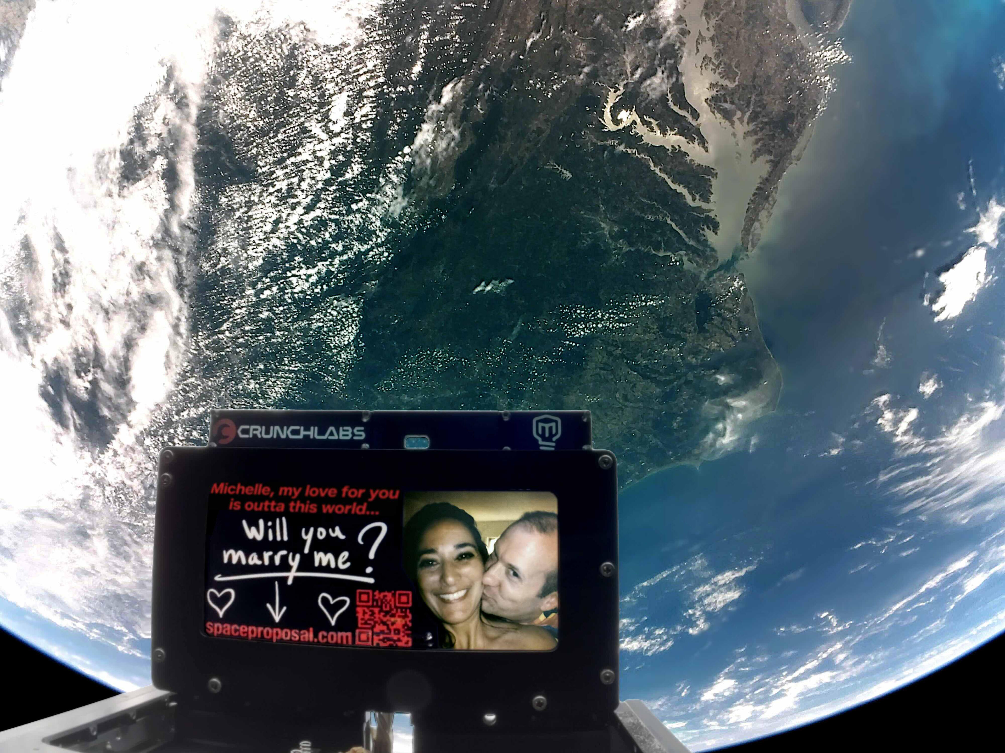 Image taken from a satellite, showing the earth in the background, and in the foreground a screen on the satellite is displaying "Michelle, my love for you is outta this world... Will you marry me?" along with a picture of Michael kissing Michelle.